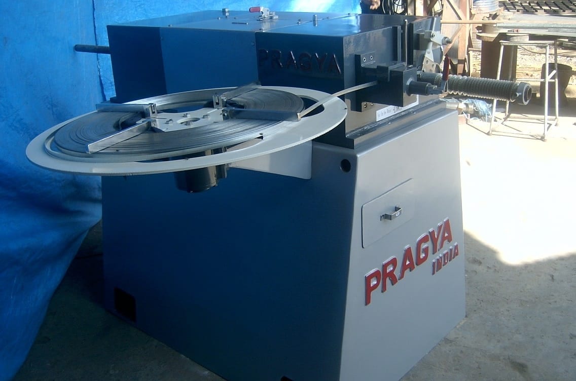 Blue and white fin tube machine lettered PRAGYA INDIA, its circular fin-strip reel mounted on top, standing in the works
