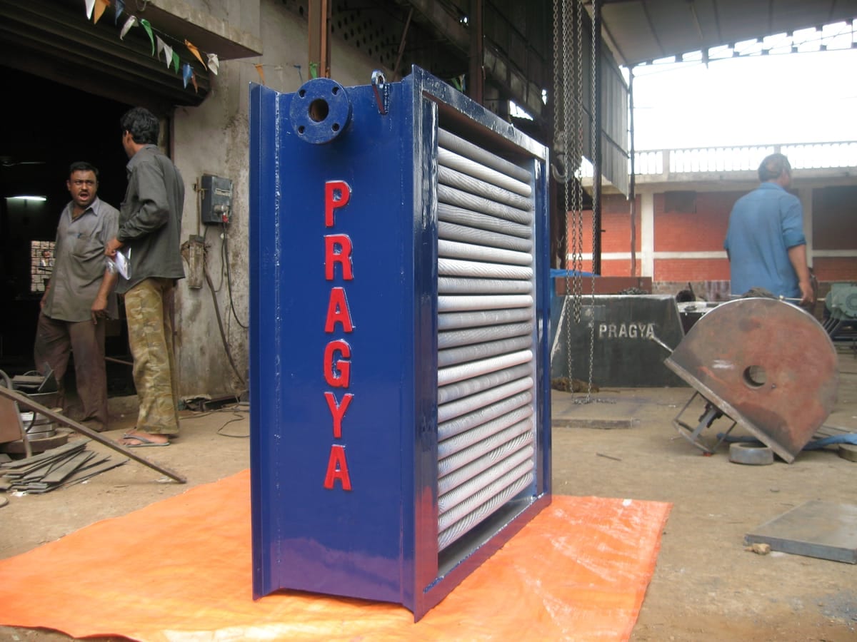Tall blue steam radiator lettered PRAGYA, its finned tube bank visible through the open face, standing on a sheet in the works